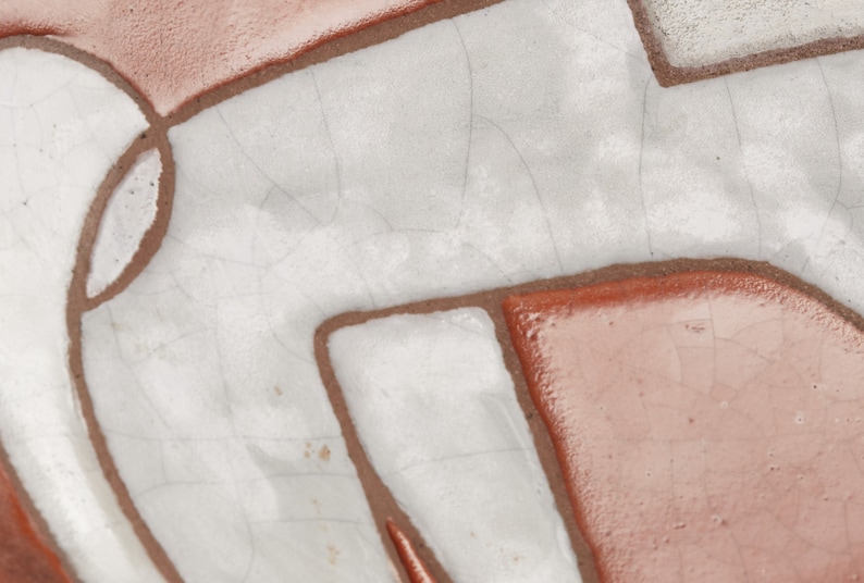 Op de afbeelding: Close-up van een keramische tegel met een witte glazuur en bruine lijnen die een geometrisch patroon vormen.