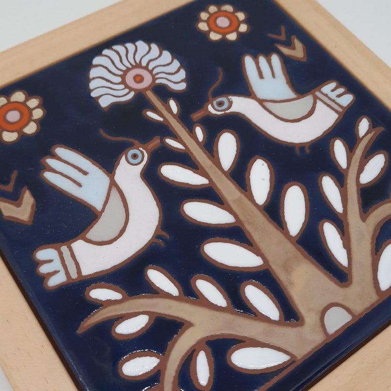 May include: A blue ceramic tile with a white floral design and two white birds. The birds are stylized and have a folk art aesthetic. The tile has a brown border.