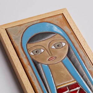 May include: A framed ceramic tile with a stylized portrait of a woman wearing a blue headscarf. The woman has brown skin, large eyes, and red lips. The tile is framed in a light wood frame.