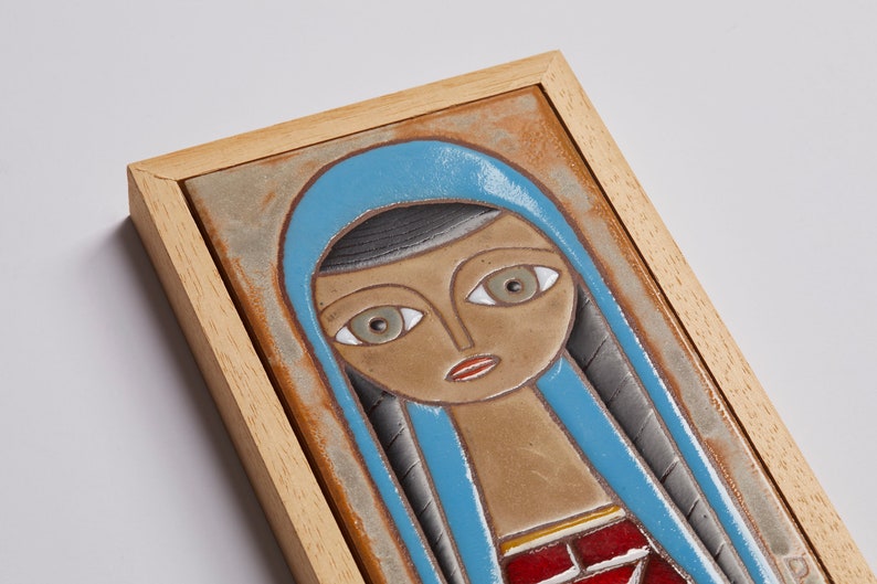 May include: A framed ceramic tile with a stylized portrait of a woman wearing a blue headscarf. The woman has brown skin, large eyes, and red lips. The tile is framed in a light wood frame.