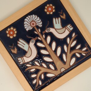 May include: A blue and white ceramic tile with a floral design and two birds. The birds are white with pink accents and are perched on a branch with white leaves. The tile has a brown wooden frame.