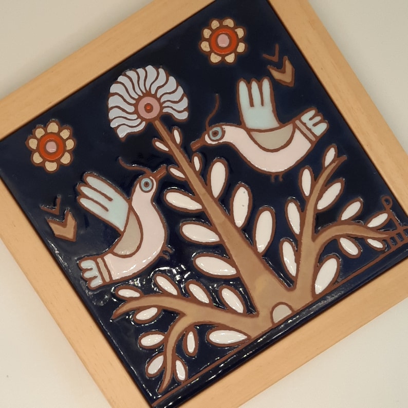 May include: A blue and white ceramic tile with a floral design and two birds. The birds are white with pink accents and are perched on a branch with white leaves. The tile has a brown wooden frame.