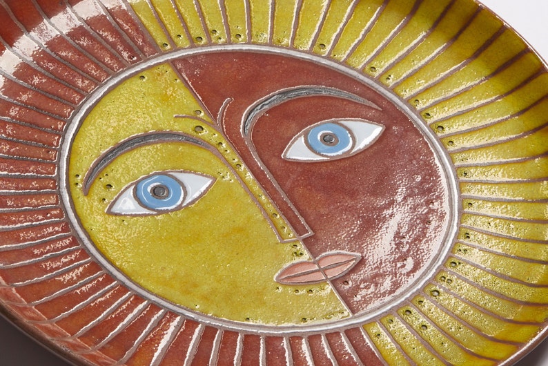 May include: A ceramic plate with a stylized sun face design. The sun has a yellow and red face with blue eyes and a pink mouth. The plate is decorated with a red and yellow sunburst pattern.