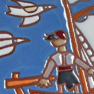 Peut inclure: Un carreau de c&eacute;ramique peint &agrave; la main repr&eacute;sentant un marin sur un bateau avec deux oiseaux blancs volant au-dessus. Le carreau a un fond bleu avec des accents orange, blanc et rouge.
