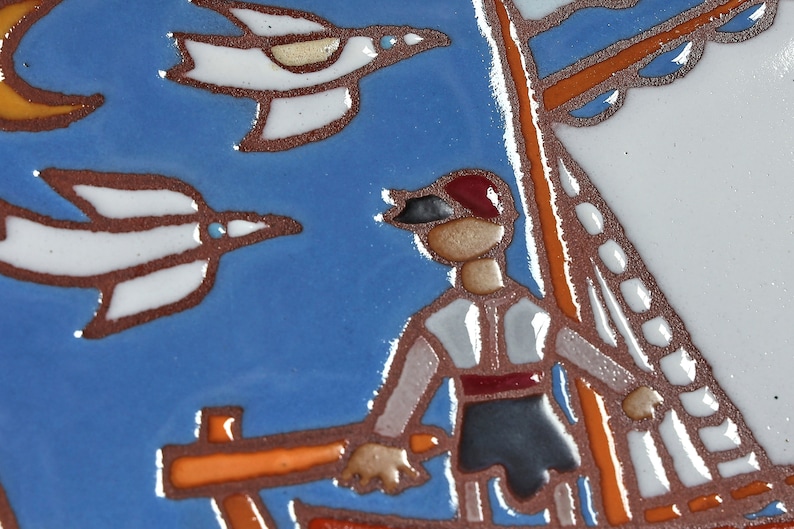 Peut inclure: Un carreau de c&eacute;ramique peint &agrave; la main repr&eacute;sentant un marin sur un bateau avec deux oiseaux blancs volant au-dessus. Le carreau a un fond bleu avec des accents orange, blanc et rouge.