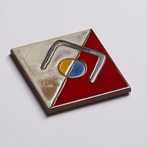 K&ouml;nnte beinhalten: Eine quadratische Keramikfliese mit einem geometrischen Design. Die Fliese ist in zwei Abschnitte unterteilt, einen wei&szlig;en und einen roten. Der wei&szlig;e Abschnitt zeigt ein silberfarbenes Design aus zwei geschwungenen Linien, die eine Form bilden, die an den Buchstaben 'U' erinnert. Der rote Abschnitt zeigt einen gelben und blauen Kreis mit einem silberfarbenen Umriss.