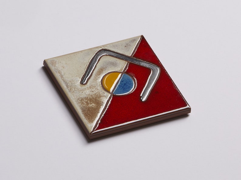 K&ouml;nnte beinhalten: Eine quadratische Keramikfliese mit einem geometrischen Design. Die Fliese ist in zwei Abschnitte unterteilt, einen wei&szlig;en und einen roten. Der wei&szlig;e Abschnitt zeigt ein silberfarbenes Design aus zwei geschwungenen Linien, die eine Form bilden, die an den Buchstaben 'U' erinnert. Der rote Abschnitt zeigt einen gelben und blauen Kreis mit einem silberfarbenen Umriss.