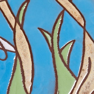 Puede incluir: Primer plano de un azulejo de cer&aacute;mica con fondo azul y un dise&ntilde;o de hojas verdes y marrones. Las hojas est&aacute;n delineadas en marr&oacute;n y tienen un centro blanco.