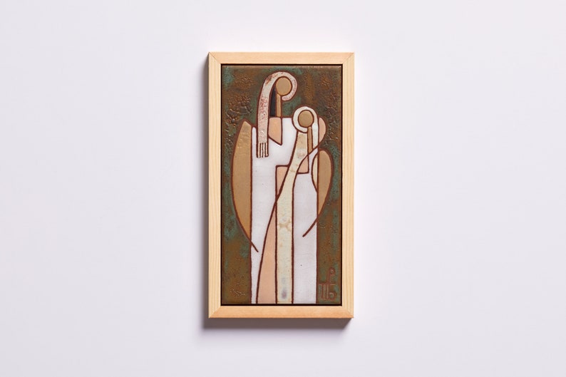 May include: A framed ceramic tile with a stylized image of two figures in white, pink, and brown against a green background. The figures are in a close embrace, with the smaller figure's head resting on the larger figure's shoulder. The tile is framed in a natural wood frame.