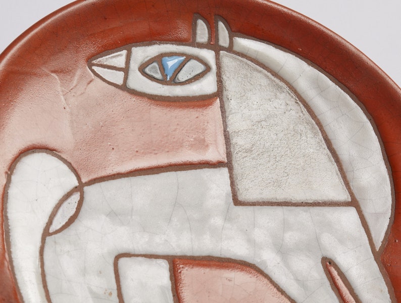Op de afbeelding: Een keramisch bord met een gestileerd wit paard ontwerp op een rode achtergrond. Het oog van het paard is blauw.