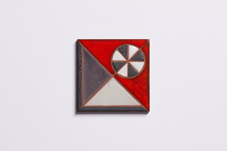 May include: A square ceramic tile with a geometric design. The tile is divided into four sections, each with a different color: red, white, black, and gray. The center of the tile features a black and white wheel design.