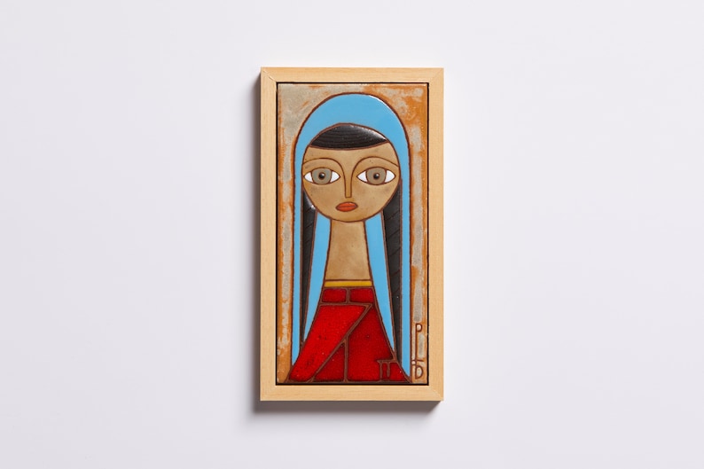 May include: A framed ceramic tile with a painting of a woman with long black hair, wearing a blue headscarf and a red dress. The woman's eyes are large and brown, and her lips are red.