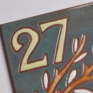 May include: A ceramic tile with the number 27 in yellow paint on a blue background. The tile has a brown branch with white leaves and red pomegranates.