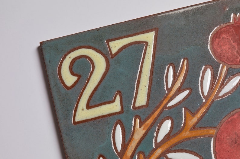 May include: A ceramic tile with the number 27 in yellow paint on a blue background. The tile has a brown branch with white leaves and red pomegranates.