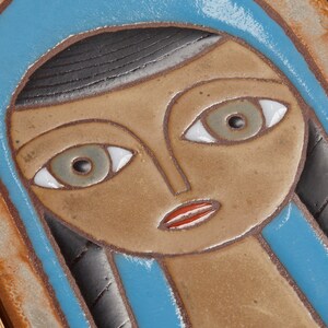 May include: A ceramic tile with a stylized portrait of a woman with blue, brown, and white details. The woman is wearing a blue headscarf and has a brown face with white eyes and a red mouth.