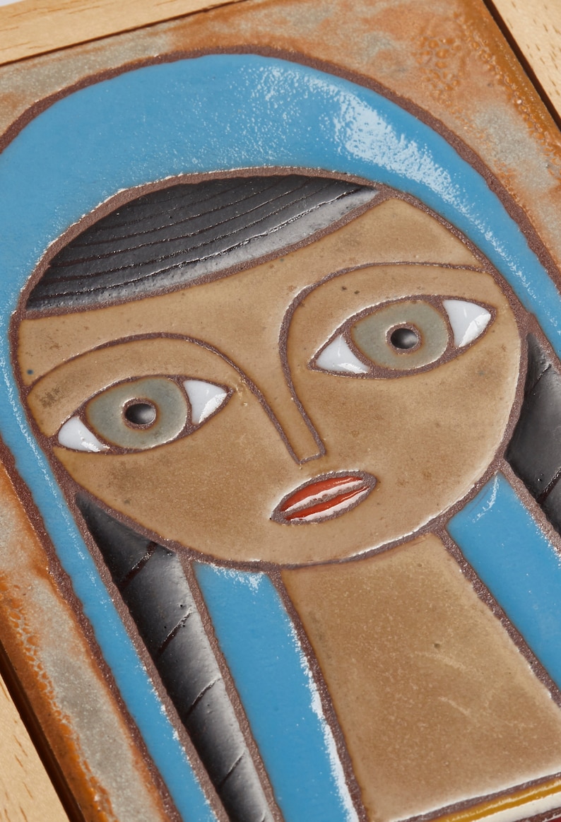 May include: A ceramic tile with a stylized portrait of a woman with blue, brown, and white details. The woman is wearing a blue headscarf and has a brown face with white eyes and a red mouth.