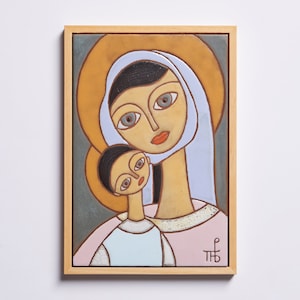 May include: A framed ceramic tile depicting a stylised image of the Virgin Mary and baby Jesus. The image is in a muted colour palette of blue, pink, yellow, and brown. The tile is framed in a light wood frame.