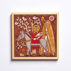 May include: A framed ceramic tile depicting a religious scene. A woman in a red dress holds a baby while riding a donkey. A man with a staff walks alongside. The background features a tree, birds, and a sun.