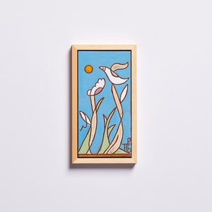 Puede incluir: Una obra de arte enmarcada que presenta un p&aacute;jaro estilizado volando sobre un campo de flores. El fondo es un cielo azul claro con un sol amarillo. La obra de arte est&aacute; en un marco de madera claro.