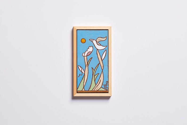 Puede incluir: Una obra de arte enmarcada que presenta un p&aacute;jaro estilizado volando sobre un campo de flores. El fondo es un cielo azul claro con un sol amarillo. La obra de arte est&aacute; en un marco de madera claro.
