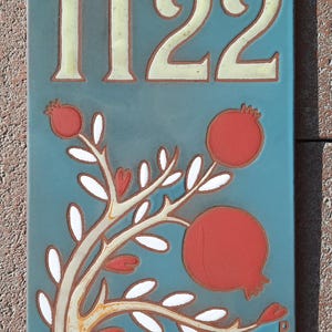 May include: A blue ceramic tile with the number 1122 in white. The tile features a stylized tree with white leaves and red pomegranates.