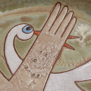 May include: A ceramic plate with a hand-painted design of a white bird with a blue eye and orange beak. The bird is depicted with its wing raised, and the background is a mottled green and brown glaze.