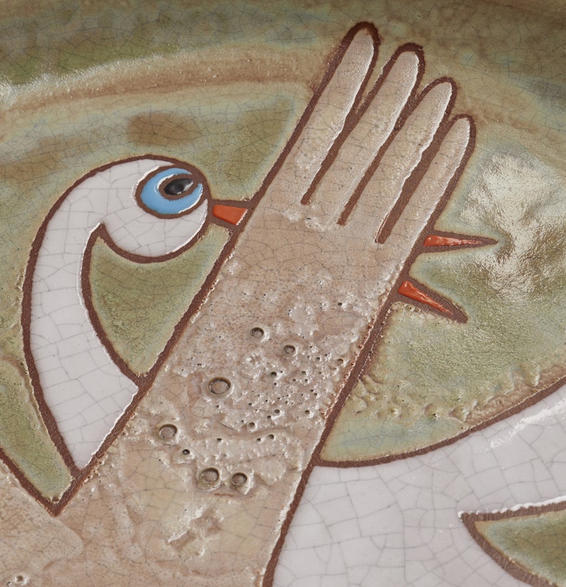 May include: A ceramic plate with a hand-painted design of a white bird with a blue eye and orange beak. The bird is depicted with its wing raised, and the background is a mottled green and brown glaze.