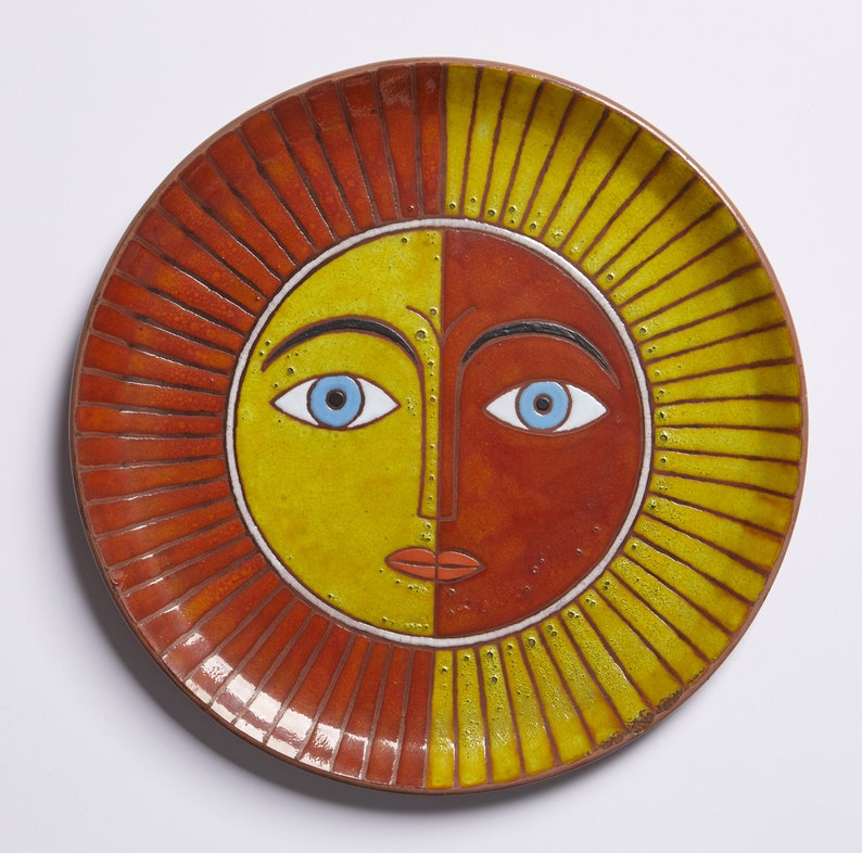 May include: A ceramic plate with a sun face design. The sun has a yellow and red face with blue eyes and a red mouth. The sun is surrounded by red and yellow rays.