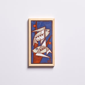 May include: A framed ceramic tile with a blue background and two white fish with orange accents. The fish are stylized and geometric in design.