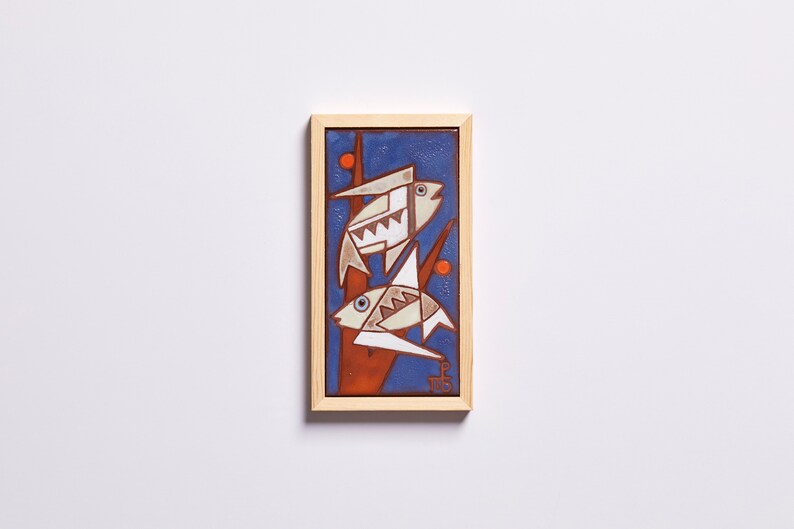 May include: A framed ceramic tile with a blue background and two white fish with orange accents. The fish are stylized and geometric in design.