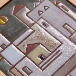 以下が含まれることがあります： 白、茶色、赤、黄色の色合いで作られた建物の幾何学模様が描かれた、額縁付きのセラミックタイル。背景は濃い緑色で、黄色の円が描かれている。