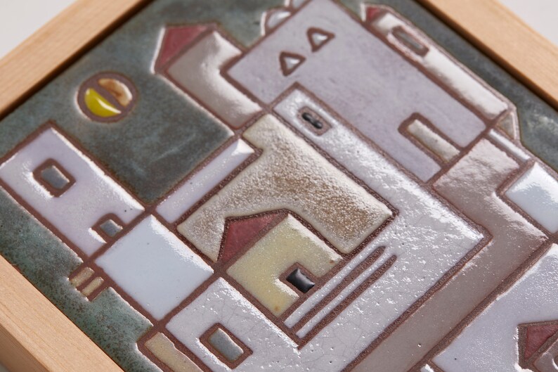以下が含まれることがあります： 白、茶色、赤、黄色の色合いで作られた建物の幾何学模様が描かれた、額縁付きのセラミックタイル。背景は濃い緑色で、黄色の円が描かれている。