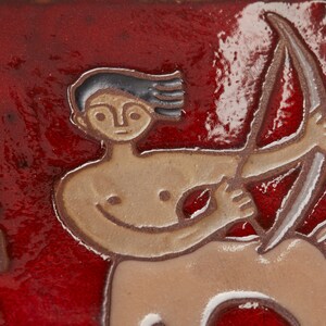 Może przedstawiać: Płytka ceramiczna z czerwonym tłem i przedstawieniem znaku zodiaku Strzelec. Znak przedstawiony jest jako centaur trzymający łuk i strzałę. Płytka ma napis "Strzelec" po lewej stronie.