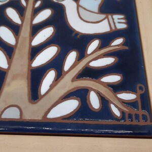 May include: A close-up of a blue and white ceramic tile with a stylized tree design. The tree has a brown trunk and white leaves. The tile is framed in wood.