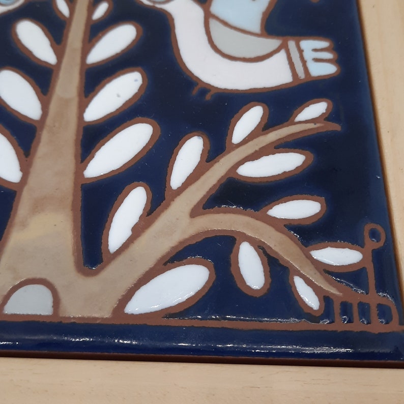 May include: A close-up of a blue and white ceramic tile with a stylized tree design. The tree has a brown trunk and white leaves. The tile is framed in wood.