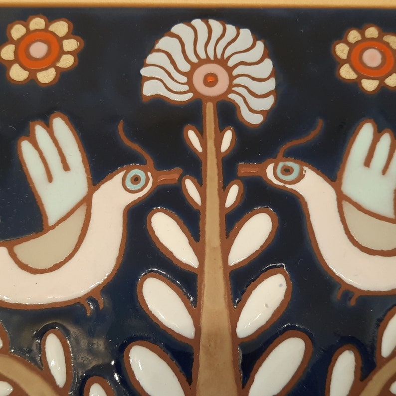 May include: A ceramic tile with a blue background featuring two white birds with blue eyes, a brown tree with white leaves, and two white flowers with pink and orange centers.