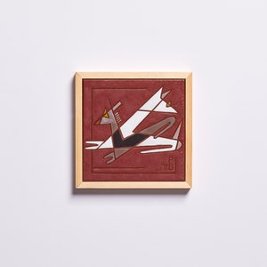 May include: A framed ceramic tile featuring a geometric design of a stylised bird in shades of white, grey, and black on a red background. The tile is framed in a light wood frame.
