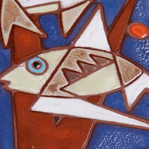 May include: A close-up of a ceramic tile with a blue background and a white fish with brown and gray details. The fish has a blue eye and is surrounded by red and brown geometric shapes.