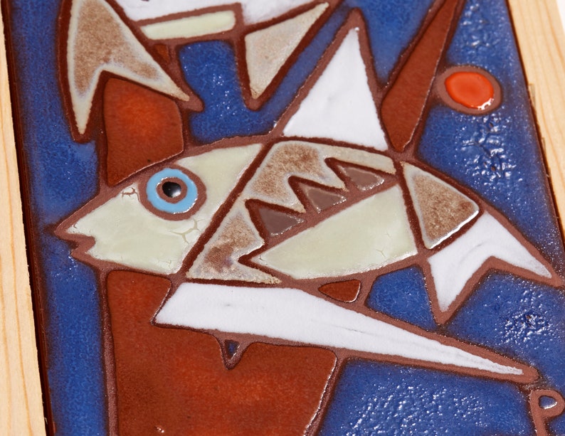 May include: A close-up of a ceramic tile with a blue background and a white fish with brown and gray details. The fish has a blue eye and is surrounded by red and brown geometric shapes.