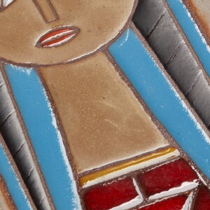 May include: A ceramic tile with a stylized image of a woman's face. The woman has blue hair, brown skin, and red lips. The tile is framed in wood.