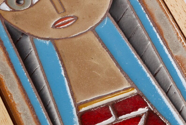 May include: A ceramic tile with a stylized image of a woman's face. The woman has blue hair, brown skin, and red lips. The tile is framed in wood.