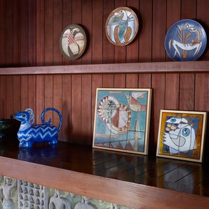 Op de afbeelding: Een houten plank met een blauw en wit keramiek dierenbeeldje, een blauwe kom en verschillende ingelijste keramische tegelkunstwerken met geometrische patronen en vogels.