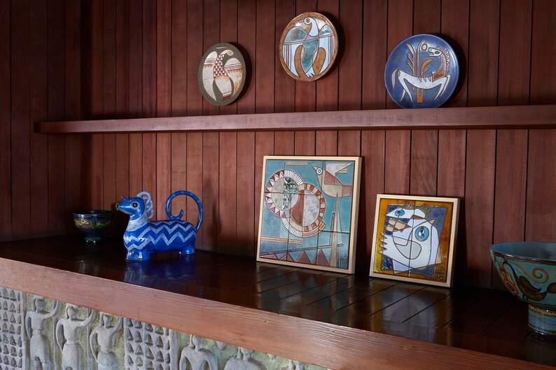 Op de afbeelding: Een houten plank met een blauw en wit keramiek dierenbeeldje, een blauwe kom en verschillende ingelijste keramische tegelkunstwerken met geometrische patronen en vogels.
