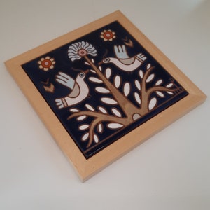 May include: A blue ceramic tile with a wooden frame features a stylized design of two birds perched on a tree branch with white leaves. The birds are white with brown accents and the tree is brown with white leaves. The background is a dark blue color.