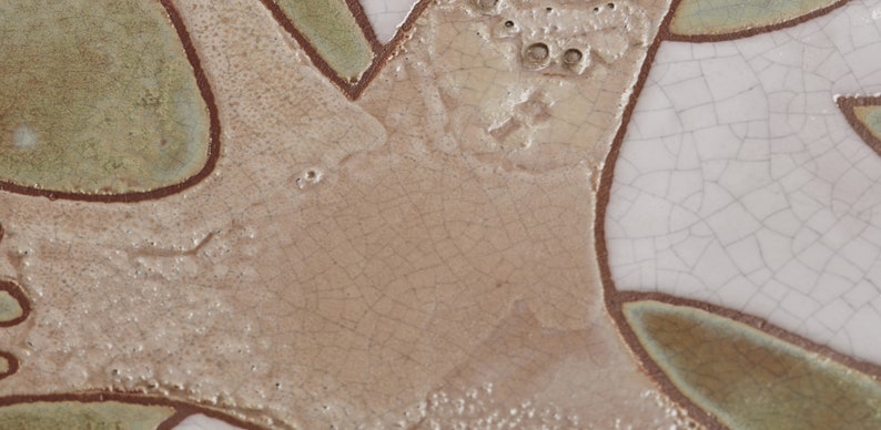 May include: Close-up of a ceramic tile with a crackled glaze. The tile has a light brown background with a pattern of green and brown lines. The lines create a design that resembles a tree branch.