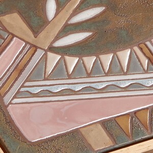 May include: A ceramic tile with a geometric design in shades of brown, pink, white, and gray. The design features a stylized animal with a tree growing from its back.