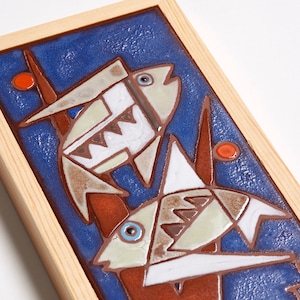 May include: A framed ceramic tile with a blue background and two stylized fish in white, brown, and orange. The fish are swimming in front of a red and brown plant.