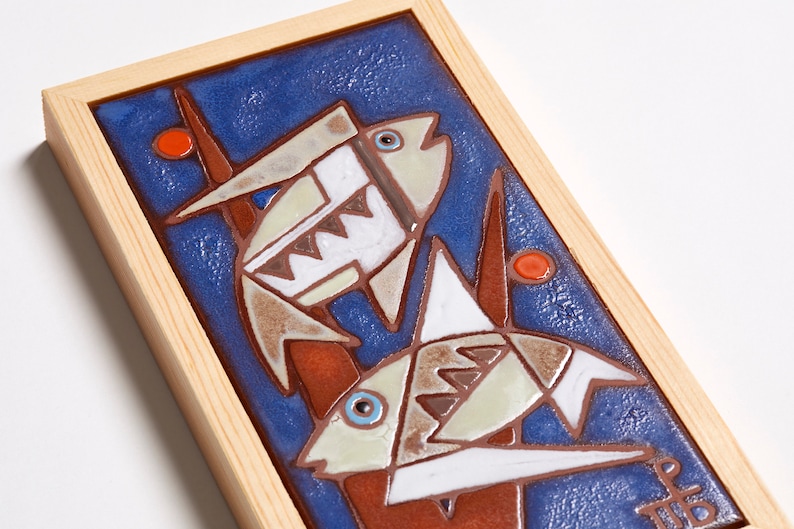 May include: A framed ceramic tile with a blue background and two stylized fish in white, brown, and orange. The fish are swimming in front of a red and brown plant.