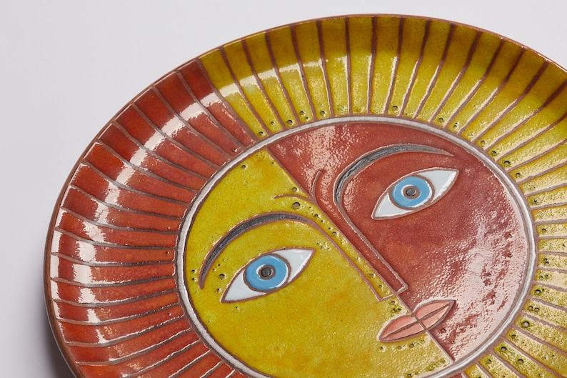 May include: A ceramic plate with a stylized sun face design. The sun has a yellow and red face with blue eyes and a pink mouth. The plate has a red and yellow rim.