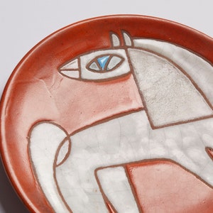 Op de afbeelding: Een rood keramiek bord met een wit gestileerd paard ontwerp. Het paard heeft een blauw oog en is omrand met bruin.
